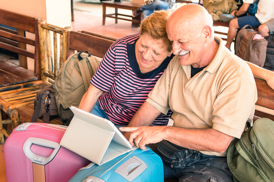 Happy senior couple sitting with digital laptop and travel baggage during adventure trip around the world - Concept of active elderly lifestyle and interaction with new trends and technologies